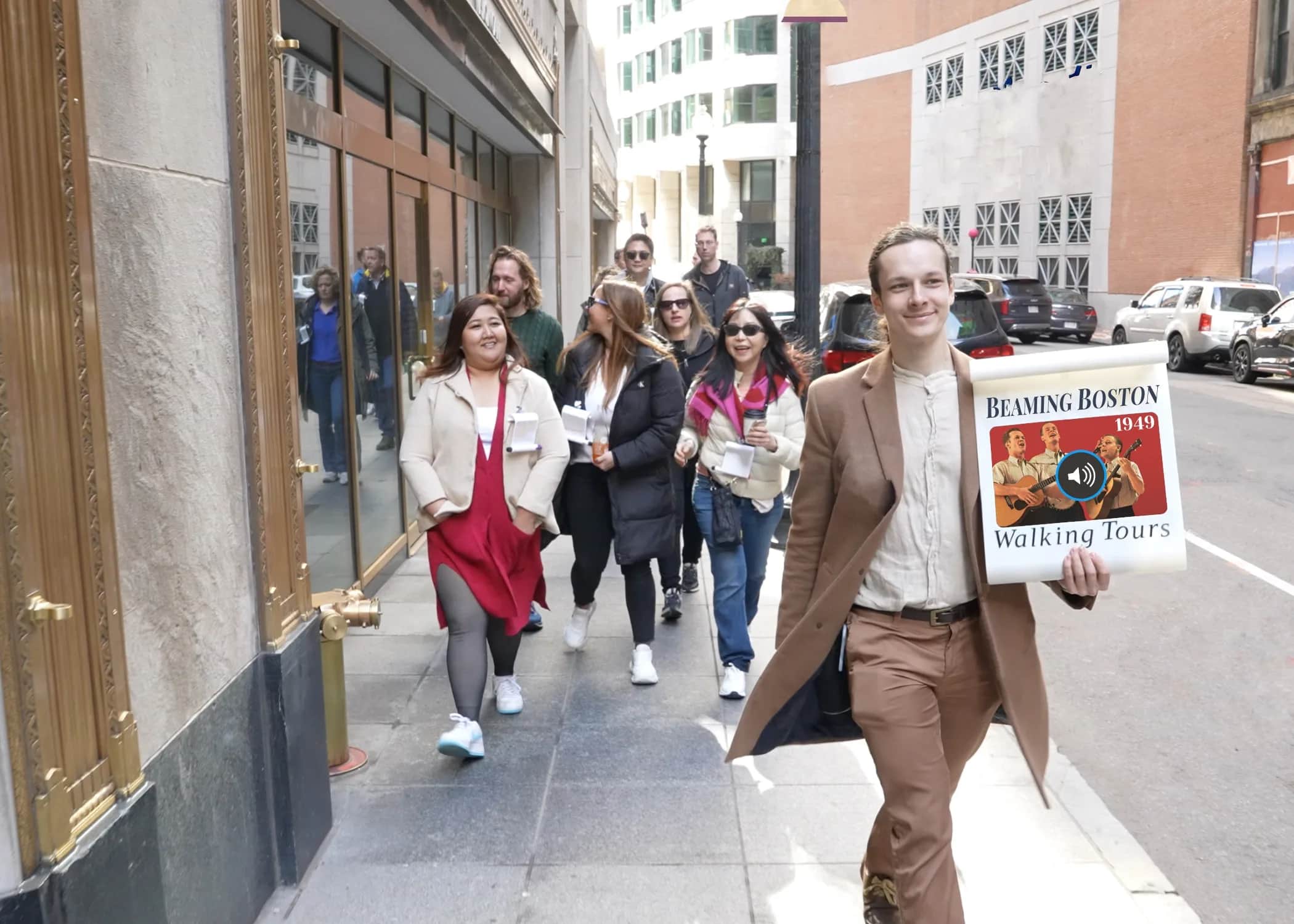 A Beaming Boston Tours guide in a camel coat walks ahead of a smiling tour group on a downtown Boston sidewalk, holding up a 'Beaming Boston Walking Tours' scroll sign